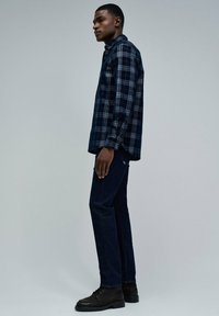Camisa de cuadros en tonos azul marino con botones y mangas largas, combinada con jeans de pierna recta en azul oscuro y botas negras con cordones. Se aprecian detalles en la textura de la tela.