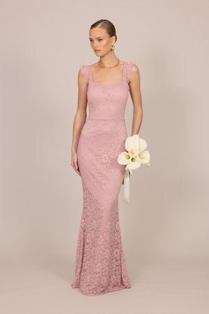 Femme en robe rose en dentelle sans manches, longue jusqu'au sol, tenant un bouquet de callas blancs avec un ruban sur un fond uni.