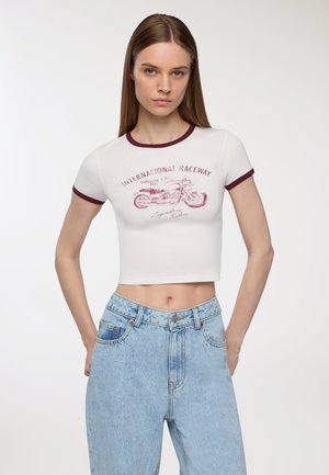 T-shirt bianca a maniche corte con bordo ribattuto bordeaux, caratterizzata da una grafica di una motocicletta e il testo "INTERNATIONAL RACEWAY". Abbinata a jeans azzurri chiari.