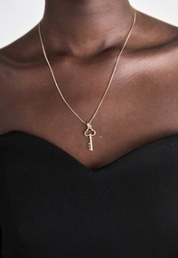 Zlatý náhrdelník s príveskom v tvare kľúča a vrchnou časťou v tvare ďateliny, ležiaci na holý pokožke nad čiernym oblečením s výstrihom do V.