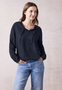 Zwarte lichte hoodie met een v-hals en trekkoord, gecombineerd met blauwe spijkerbroek. De stof heeft een subtiel gestructureerd patroon.