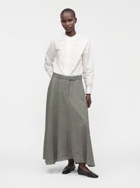 Femme debout portant un chemisier blanc à manches longues, une jupe grise longueur cheville et des chaussures plates noires sur un fond clair uni.