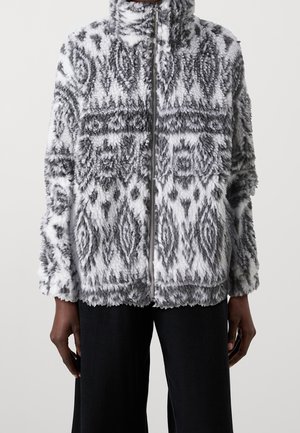 Henkilö, joka on pukeutunut harmaan ja valkoisen kirjavan pehmeän vetoketjullisen takin, jossa korkea kaulus, ja mustiin housuihin. Henkilö seisoo yksiväristä taustaa vasten.