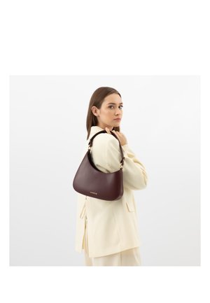 Donna in completo crema che tiene una borsa a tracolla marrone scuro con dettagli in metallo color oro, in piedi su uno sfondo bianco semplice.
