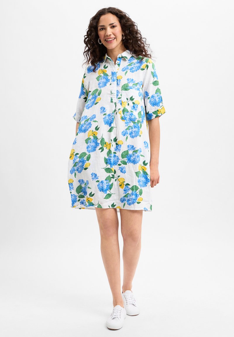 Blumenhemdkleid in Weiß mit Druck von blauen Hortensien und Zitronen, kurzen Ärmeln, Kragen und einer Brusttasche. Kombiniert mit weißen Sneakern.