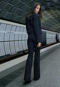 Crni odijelo sa prugicama s uskim sakoom i širom hlačama, teksturirana tkanina, nosi se s crnim visokim potpeticama, smješteno u modernoj podzemnoj željeznici.