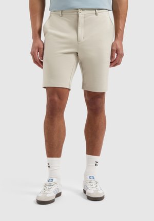 Light beige shorts with a smooth texture, featuring a front zipper and button closure, paired with white sports socks and casual sneakers.