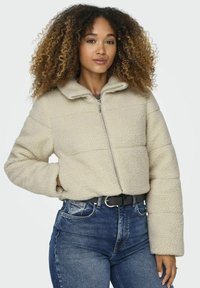 Beige cropped fleece jack met ritssluiting en een opstaande kraag. Gelaagd, pluizig materiaal; gecombineerd met slim-fit blauwe jeans en een zwarte riem.