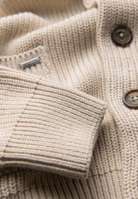 Beige gestrickter Pullover mit gerippter Textur, flachem Kragen und braunen Knöpfen mit erhabenen Details. Enthält ein Stoffetikett.