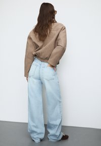 Rjav skrajšan pulover, svetlo modri hlače širokega kroja z visokim pasom in rjave čevlje, prikazani s hrbta na enobarvni podlagi.