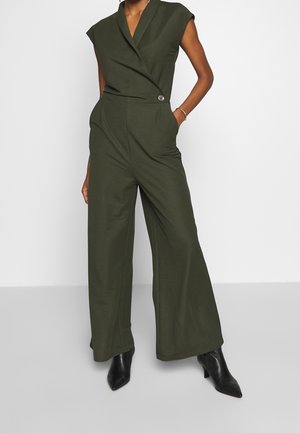 Femme portant une combinaison sans manches vert olive à jambes larges et des bottines noires à bout pointu, debout avec une main dans la poche.