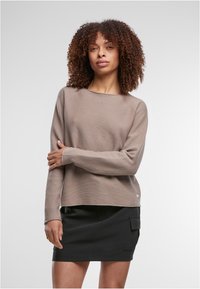 Göndörhajú nő, aki taupe színű hosszú ujjú pulóvert és fekete oldalzsebes szoknyát visel, egyszínű világos háttér előtt áll.
