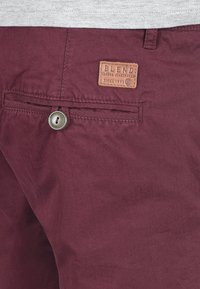 Bordo hlače s glatkom teksturom, sa zakrpanom etiketom koja glasi "BLEND" i vidljivim metalnim dugmetom na stražnjem džepu.