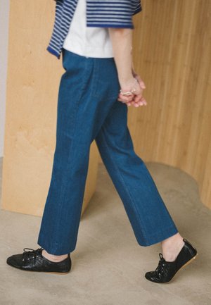 Persoon die binnenshuis loopt, gekleed in blauwe wijde broek, zwarte veterlaarzen, een gestreept blauw-wit shirt, met de handen achter de rug gekruist.