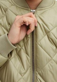 Licht olijfgroene geborduurde bomberjack met ribgebreide manchetten en kraag, voorzien van een voorrits. Hand die de ritstrekkertje vasthoudt.