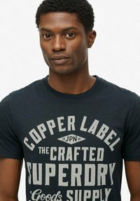 Mand iført en marineblå t-shirt med hvid tekst, der står "COPPER LABEL THE CRAFTED SUPERDRY Goods SUPPLY" på en lys ensfarvet baggrund.