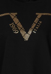 Černá látka se zlatým geometrickým vzorem ve tvaru "V", s rokem 1969 a slovem "ITALIA" v zlatém písmu.