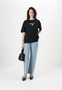 Black cotton t-shirt with white graphic text, paired with light blue jeans featuring silver embellishments and a black textured handbag.