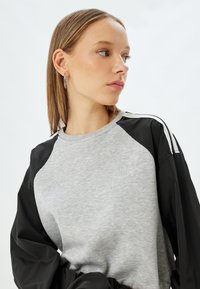 Grijze sweatshirt met zwarte lange mouwen met een glanzende afwerking en witte accentstrepen op de schouders. Zachte textuur met een relaxte pasvorm.