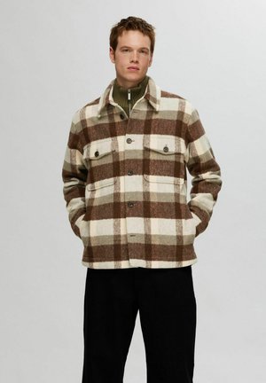 Young man wearing a brown and cream checkered wool jacket over a green zip-up sweater with hands in pockets, standing against a plain background.