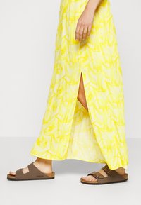 Sandales Birkenstock brunes avec deux brides réglables, portées avec une maxi robe fleurie jaune à fente sur le côté, sur un fond blanc.