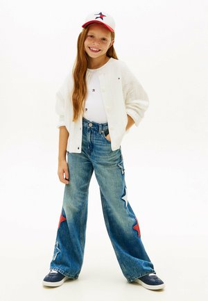 Jeune fille souriante aux longs cheveux roux porte une casquette blanche à motifs d'étoiles, un cardigan blanc, un haut blanc, un jean pattes larges avec des patchs étoiles rouges et blanches, et des baskets bleu marine.
