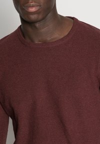 Bordeaux gestrickter Pullover mit Rundhalsausschnitt, strukturiertem Stoff und mittlerem Gewicht. Nahaufnahme hebt das Muster und die Farbreichtum hervor.