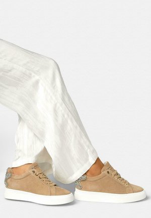 Feet wearing beige suede sneakers with rhinestone detail on heel, paired with loose white textured pants against a plain background.