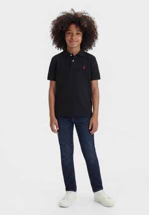 Berniukas su garbanotais plaukais, vilkintis juodą polo marškinėlį, tamsiai mėlynas džinsas ir baltus sportinius batelius, stovintis atsipalaidavusio pozu žiūrėdamas į priekį.