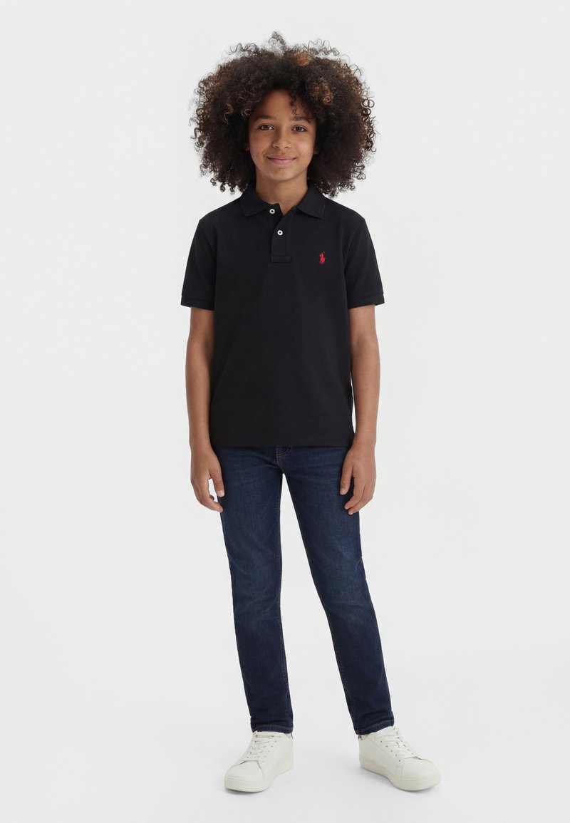 Garçon aux cheveux bouclés portant un polo noir, un jean bleu foncé et des baskets blanches, debout face à l'avant avec une posture détendue.