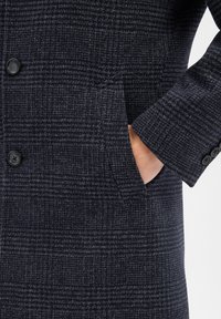 Marineblauer Karottenmantel aus strukturiertem Stoff, mit einem Ein-Knopf-Verschluss und Seitentaschen. Enthält schwarze Knöpfe an den Ärmeln.