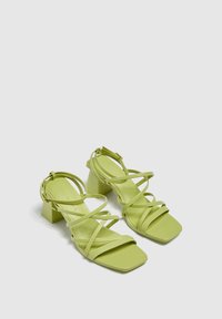 Un paio di sandali strappy verde lime con tacco a blocco e punta quadrata su uno sfondo chiaro e uniforme.