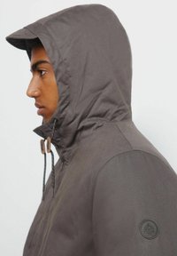 Szara kurtka z kapturem o matowym wykończeniu, z regulowanymi sznurkami i okrągłym logo na lewym rękawie. Prosty, elegancki design.