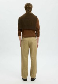 Personne aux cheveux courts châtain clair portant un pull marron avec un châle marron foncé et un pantalon beige, debout de dos sur un fond blanc.