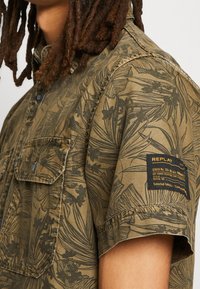 Korte mouwen shirt in olijfgroen met een zwart bloemenpatroon, met twee voorkruiken en een zwart labelpatch op de mouw.