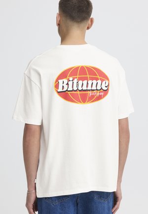 Persona che indossa una maglietta bianca con un logo del globo rosso e giallo e la scritta "Bitume skateshop" sulla schiena, abbinata a jeans blu.