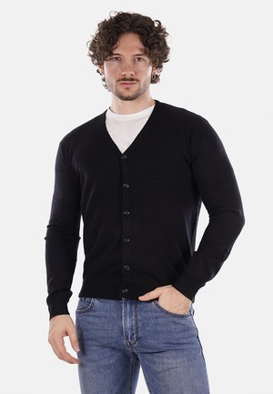 Cardigan nero realizzato in un tessuto morbido, con scollatura a V, maniche lunghe e sei bottoni frontali. Indossato sopra una camicia bianca, con un design aderente.