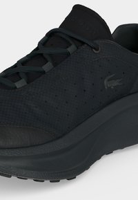 Baskets noires avec dessus texturé, détails perforés, lacets arrondis et semelle épaisse, avec un petit logo crocodile sur le côté.
