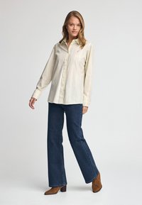 Camicia button-up beige chiaro con righe bianche verticali, indossata con jeans a zampa d'elefante blu scuro e stivali con tacco marroni, su uno sfondo neutro.