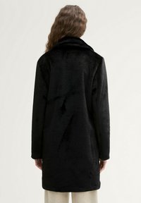 Cappotto in faux fur nero con colletto, maniche lunghe e un design strutturato dritto di media lunghezza. Texture liscia con un aspetto morbido.