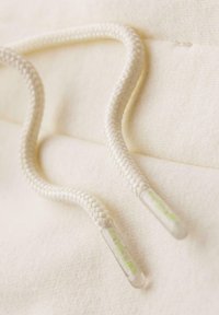 Splétané krémové tkaničky s průhlednými konci a zeleným textem. Hladký povrch a zaoblený tvar, položené na světlém textilním pozadí.