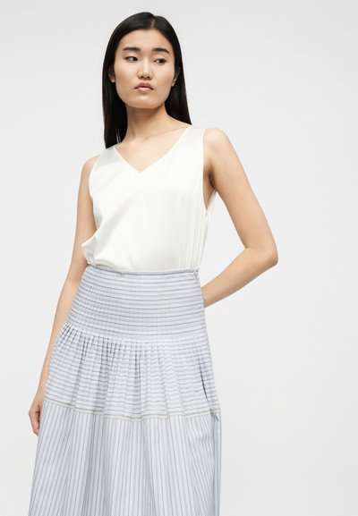 Femme aux cheveux longs et foncés portant un haut sans manches blanc à col en V et une jupe plissée taille haute avec de fines rayures horizontales et verticales.