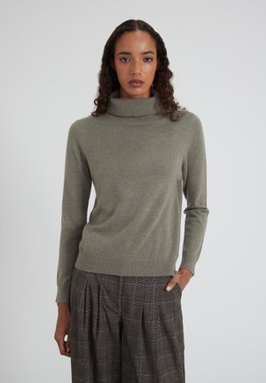 Donna con capelli ricci che indossa un maglione a collo alto grigio e pantaloni a pieghe scozzesi marroni, in piedi con una mano in tasca.