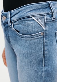 Κοντινό πλάνο ατόμου που φοράει τζιν Replay ανοιχτού μπλε χρώματος με ορατή μπροστινή τσέπη, θηλιά ζώνης και μικρή ετικέτα με το λογότυπο Replay κοντά στην τσέπη.