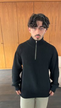 Sweater preto com nervuras, colarinho alto e fecho de meio zíper. Mangas longas com punhos justos. Corte espaçoso. Usado sobre calças de cor clara.
