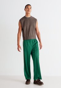 Sweater castanho sem mangas com decote redondo, combinado com umas calças verdes largas com uma faixa lateral e sapatos castanhos.