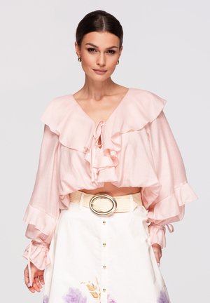 Femme aux cheveux foncés portant un chemisier rose à volants et une jupe blanche avec des détails floraux et une large ceinture, debout devant un fond uni.