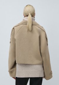 Blond inimene ponihobusega, seljas beež lühike jakk kerge kudumipusa ja mustade pükstega, nähtuna seljalt ühtlasel taustal.