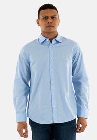 Camisa de botones azul claro en una tela suave, con mangas largas, cuello y un pequeño logo en el pecho. Combinada con jeans oscuros.