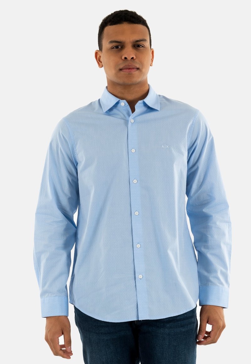 Camisa de botones azul claro en una tela suave, con mangas largas, cuello y un pequeño logo en el pecho. Combinada con jeans oscuros.